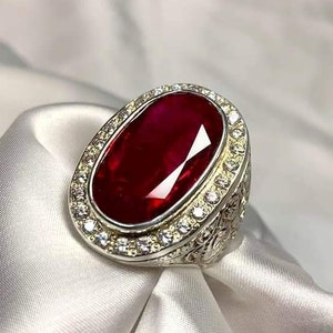 May include: A silver ring featuring a large oval red gemstone, encircled by smaller white gemstones.
