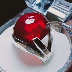 May include: A vibrant red gemstone ring set in a silver band, displayed in a clear, faceted box. The ring's gemstone is a deep, saturated red, and the silver band has a modern, angular design. The box is held by a hand.