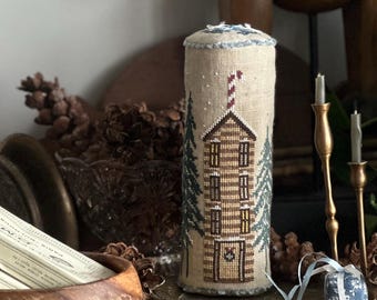 May include: A still life arrangement featuring a wooden bear figurine, a decorative cross-stitch cylinder with a house design, and a green wine bottle with a candle. A wooden bowl holds two boxes of matches.