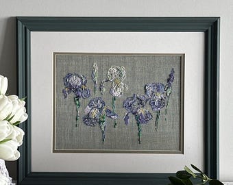 May include: A framed embroidery piece featuring five iris flowers in shades of purple, blue, and white, stitched onto a gray fabric background. The artwork is enclosed in a dark green rectangular frame with a white mat board border.
