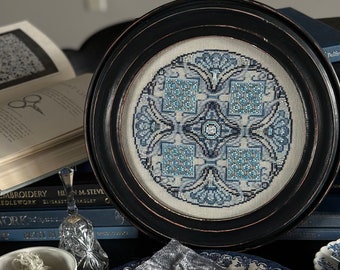 May include: A framed cross-stitch piece with a geometric blue and white design. The artwork is displayed on a table with books, a plate, and other decorative items. The frame is dark with a distressed finish.