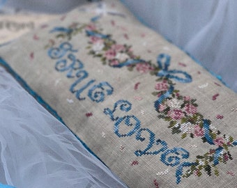 May include: A rectangular beige linen pillow featuring cross-stitched text that reads TRUE LOVE in blue, surrounded by a floral garland of pink roses and blue ribbons. The pillow has a bright blue trim and is accented with small white beads.
