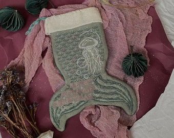May include: A handmade, mermaid tail-shaped Christmas stocking. The stocking is primarily sage green with a detailed embroidered design of a jellyfish and bubbles. The tail features pink accents and is set against a backdrop of pink fabric and dried flowers.