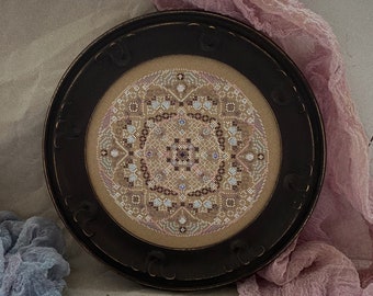 May include: A framed, circular cross-stitch design with a detailed geometric pattern in shades of beige, brown, and blue. The dark brown frame has decorative cutouts. A pink and blue fabric and vintage scissors are in the background.