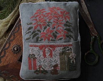 May include: A gray decorative pillow with a cross-stitched poinsettia design. The pillow features a pocket with cross-stitched gift boxes and bows. The background includes pine branches, a wooden box, and vintage scissors.