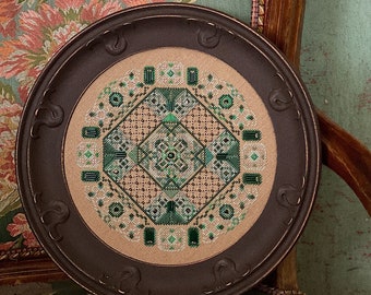 May include: A framed cross-stitch featuring a geometric design in shades of green and beige. The round, dark brown frame has decorative swirls. The piece is displayed on a floral patterned chair.