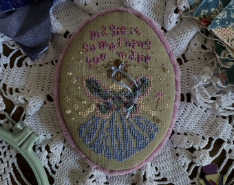 May include: An oval-shaped, embroidered piece with the text "My life is complicated you make it more". The embroidery features a butterfly and floral design in blue and pink, framed by a pink border. The piece is set on a white lace surface.