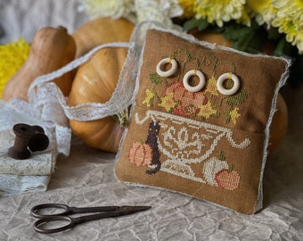 May include: A square brown fabric pincushion featuring a cross-stitched design of a black cat, pumpkins, and a decorative bowl. Three small wooden rings are attached to the top. The edges are finished with white lace trim.