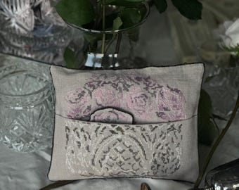 May include: A rectangular, light gray decorative pillow with a floral design. The pillow features a pocket with a detailed, silver-colored pattern and a top section with pink floral embroidery. The pillow is placed on a white surface.