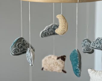 May include: A baby mobile featuring a wooden ring with hanging plush shapes. The mobile includes a beige crescent moon, fluffy clouds, a sheep, and other soft, textured elements in shades of white, blue, and beige. The mobile is designed for a nursery.