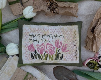 May include: A cross-stitched piece with the text "Where flowers bloom, so does hope" and pink tulips. The piece is on a green fabric. The image also includes white tulips, seed packets, and a small gardening trowel.