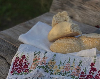 May include: A white linen towel with a decorative cross-stitch floral border in shades of red, pink, and yellow, finished with long white fringe, holding several pieces of crusty, golden-brown baked bread on its surface.