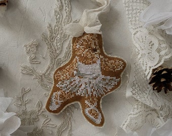 May include: A handmade, cross-stitched Christmas ornament in the shape of a reindeer. The ornament features a brown body with white and gray detailing. It is suspended by a cream-colored ribbon. The background includes lace and pinecones.