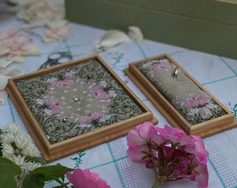 May include: Two framed needlepoint pieces with floral designs in pink and gray, displayed on a checkered tablecloth. A pink geranium and white flowers are in the foreground. A green box and a wooden tray with threads are in the background.
