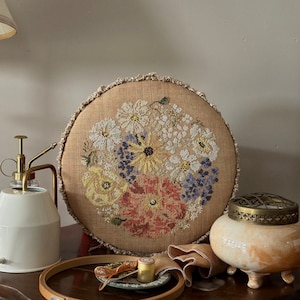 Könnte beinhalten: Runder, beigefarbener Kreuzstich mit Blumenmuster auf leinenartigem Stoff. Die Stickerei zeigt Blumen in Weiß, Gelb, Rot und Blau. Ein hölzerner Stickrahmen, ein kleiner Teller mit Faden und ein Wassersprüher sind ebenfalls zu sehen.