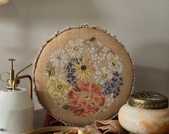 May include: A round, beige cross-stitch floral design on a linen-like fabric. The embroidery features a variety of flowers in shades of white, yellow, red, and blue. A wooden embroidery hoop, a small plate with thread, and a water mister are also visible.