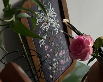 May include: A rectangular wooden frame containing a dark gray fabric panel with an intricate floral embroidery design in white, pink, and green thread. The embroidery depicts a tall, blooming plant with delicate petals and detailed foliage.