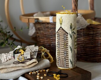 May include: A cylindrical pin cushion with a cross-stitched house and corn design, resting on a wooden board. The scene includes embroidery floss, fabric, scissors, and a basket, suggesting a crafting theme. The color palette is neutral with pops of yellow.