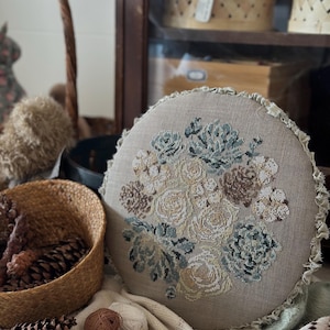 Puede incluir: Un cojín decorativo redondo beige con un diseño floral de punto de cruz en tonos crema, marrón y verde azulado. El cojín tiene un borde con volantes y se muestra sobre una manta blanca texturizada. Una cesta de piñas y ovillos de lana están en primer plano.