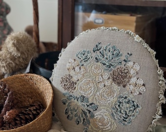 May include: A round, beige decorative pillow with a floral cross-stitch design in shades of cream, brown, and teal. The pillow has a ruffled edge and is displayed on a textured white blanket. A basket of pinecones and yarn balls are in the foreground.