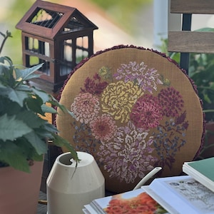 Op de afbeelding: Een rond mosterdkleurig kussen met een kruissteek bloemenontwerp in roze, gele en paarse tinten. Het kussen heeft een donkerrode rand. Een klein houten decoratief huisje en een witte gieter staan op de voorgrond.