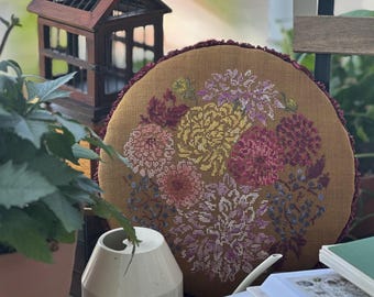 May include: A round, mustard-colored pillow with a cross-stitched floral design in shades of pink, yellow, and purple. The pillow has a dark red trim. A small wooden decorative house and a white watering can are in the foreground.