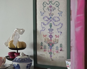 May include: A framed cross-stitch sampler with a green frame. The sampler features a symmetrical design with bows, floral elements, and a central candelabra motif. The color palette includes pink, purple, green, and blue threads. The sampler is displayed on a wooden surface.
