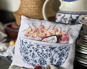 May include: A rectangular linen pillow featuring a cross-stitch design of a blue and white patterned bowl filled with yellow and red cherries. The pillow has a dark blue stitched edge and a small pocket containing a matching cherry-shaped ornament.