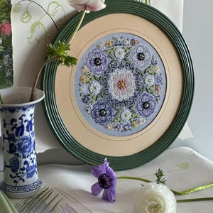 Può includere: Un ricamo circolare incorniciato con un motivo floreale nei toni del viola, bianco e giallo. Il ricamo è posto su un tessuto azzurro e incorniciato in una cornice rotonda verde. Un vaso blu e bianco e dei fiori sono in primo piano.
