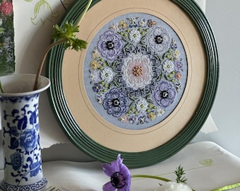 May include: A framed, circular embroidery with a floral design in shades of purple, white, and yellow. The embroidery is set against a light blue fabric and framed in a green, round frame. A blue and white vase and flowers are in the foreground.