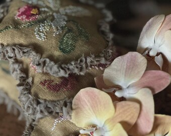 May include: Close-up of a stack of embroidered fabric squares with a floral design in pink, green, and white on a tan background. The edges are trimmed with a gray, ruffled fabric. Delicate peach and yellow orchids are in the foreground.