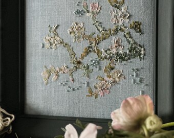 May include: A framed cross-stitch embroidery featuring a floral design with pink and cream-colored blossoms and green leaves. The embroidery is set against a light blue fabric background. The frame is dark green.