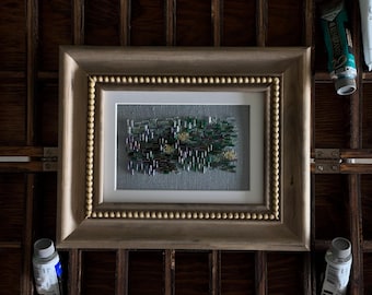 May include: A rectangular framed embroidery piece featuring a textured abstract design with vertical green, purple, and yellow thread accents on a gray fabric background, enclosed in a wooden frame with a beaded inner border and a white mat.