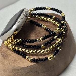 May include: A silver smartwatch with a gold and black beaded band. The band is made of small, round beads in alternating gold and black colors. The watch is resting on a wooden surface.
