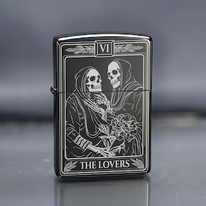 May include: Black metal lighter with a tarot card design. The image features two skeletal figures in hooded cloaks, holding a flower. The text "VI" is at the top, and "THE LOVERS" is at the bottom. The lighter has a glossy finish.