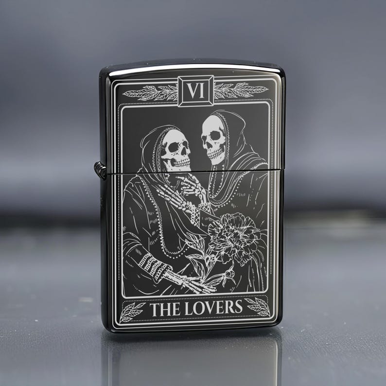 May include: Black metal lighter with a tarot card design. The image features two skeletal figures in hooded cloaks, holding a flower. The text "VI" is at the top, and "THE LOVERS" is at the bottom. The lighter has a glossy finish.