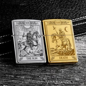 May include: Two metal lighters, one silver and one gold, each featuring tarot card designs. The silver lighter displays "THE SUN" with a skeleton on horseback. The gold lighter shows "DEATH" with a skeleton on horseback. Both have a rectangular shape.