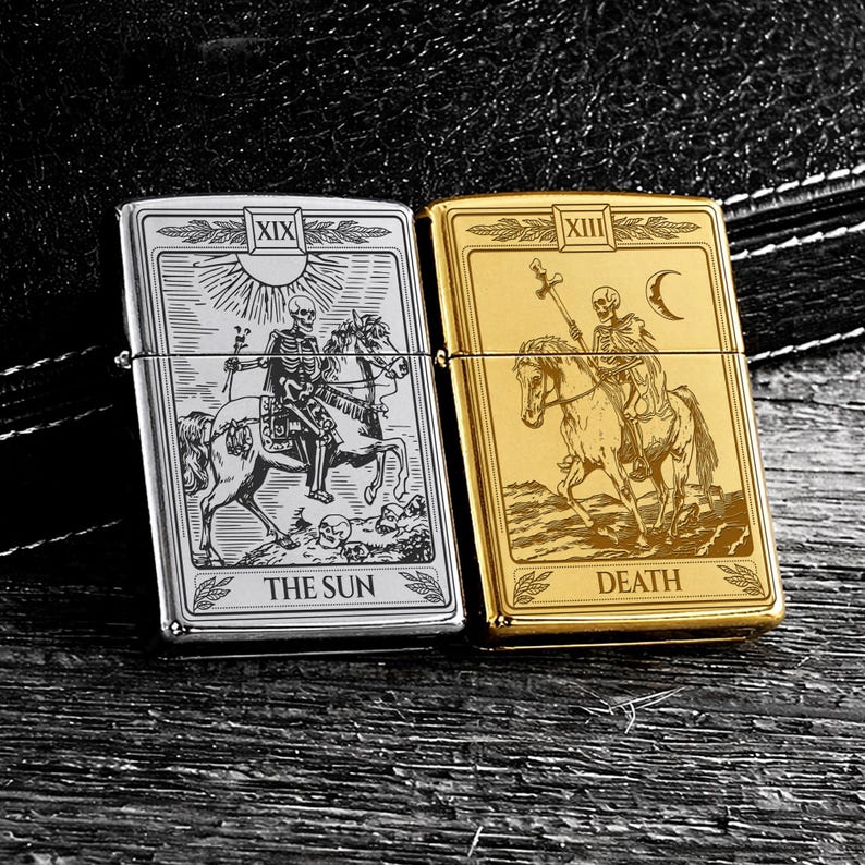 May include: Two metal lighters, one silver and one gold, each featuring tarot card designs. The silver lighter displays "THE SUN" with a skeleton on horseback. The gold lighter shows "DEATH" with a skeleton on horseback. Both have a rectangular shape.