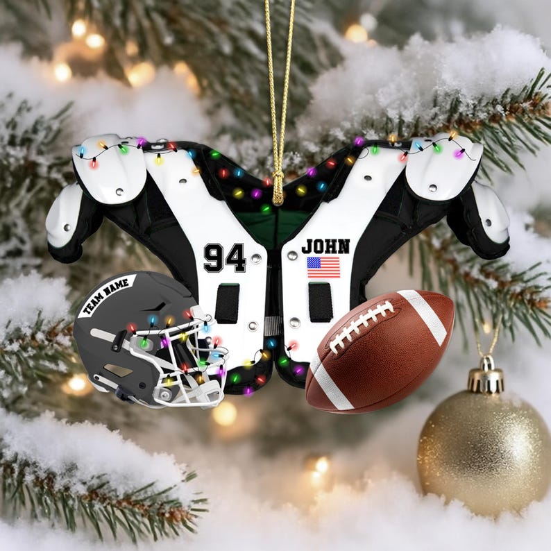 K&ouml;nnte beinhalten: Ein weihnachtlicher Football-Schmuck mit Schulterpolstern, einem Helm und einem Football. Die Schulterpolster sind wei&szlig; und schwarz mit der Nummer 94 und dem Namen "JOHN" mit einer amerikanischen Flagge. Der graue Helm tr&auml;gt den Schriftzug "TEAM NAME".