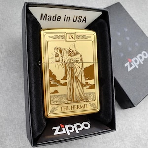 May include: Gold-colored Zippo lighter featuring an engraved image of The Hermit tarot card. The lighter is in a black box with the text "Made in USA" and the Zippo logo. The Hermit is depicted holding a lantern.