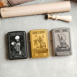 May include: Three metal lighters with tarot card designs. The lighters feature the Strength, Death, and The Magician cards. The Strength lighter is black, the Death lighter is gold, and The Magician lighter is silver. A rolled parchment and wax seal are in the background.
