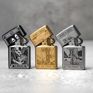 May include: Three metal lighters with tarot card designs. The lighters are black, gold, and silver. Each lighter has a different tarot card design: "The Lovers", "The Tower", and "The Hermit". The lighters are open, revealing the internal mechanism.