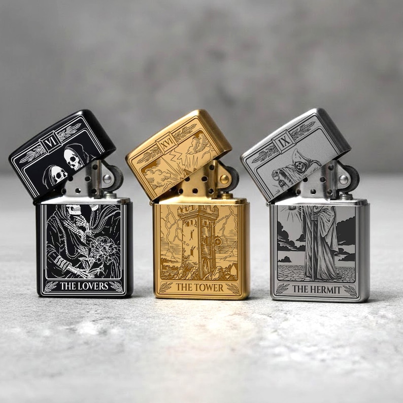 May include: Three metal lighters with tarot card designs. The lighters are black, gold, and silver. Each lighter has a different tarot card design: "The Lovers", "The Tower", and "The Hermit". The lighters are open, revealing the internal mechanism.