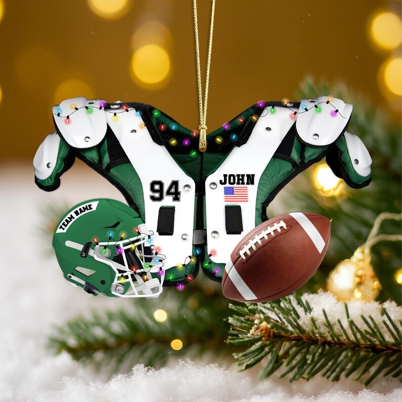 K&ouml;nnte beinhalten: Fu&szlig;ball-Weihnachtsschmuck mit einem gr&uuml;n-wei&szlig;en Schulterpolster-Design mit der Nummer 94 und dem Namen "JOHN". Enth&auml;lt einen braunen Football und einen gr&uuml;nen Helm. Der Schmuck ist mit kleinen, bunten Lichtern verziert.