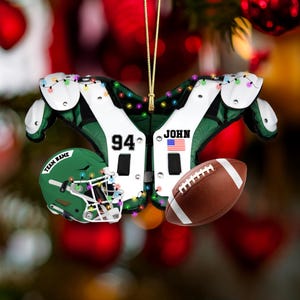 K&ouml;nnte beinhalten: Ein weihnachtlicher Football-Ornament mit gr&uuml;n-wei&szlig;en Schulterpolstern, einem gr&uuml;nen Helm mit der Aufschrift "TEAM NAME" und einem braunen Football. Das Ornament ist mit kleinen, bunten Lichtern geschm&uuml;ckt und tr&auml;gt den Namen "JOHN" und die Nummer "94".