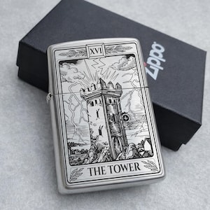 May include: A silver Zippo lighter with a detailed engraving of The Tower tarot card. The design features a tower struck by lightning, with the text "THE TOWER" at the bottom. The lighter is next to a black box.
