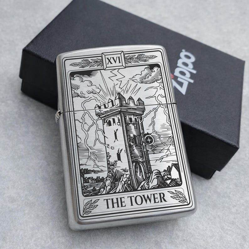 May include: A silver Zippo lighter with a detailed engraving of The Tower tarot card. The design features a tower struck by lightning, with the text "THE TOWER" at the bottom. The lighter is next to a black box.