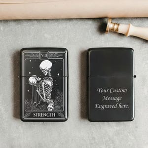May include: Black metal lighter with a tarot card design featuring a skeleton figure and the word "STRENGTH". The other lighter is black with the text "Your Custom Message Engraved here."