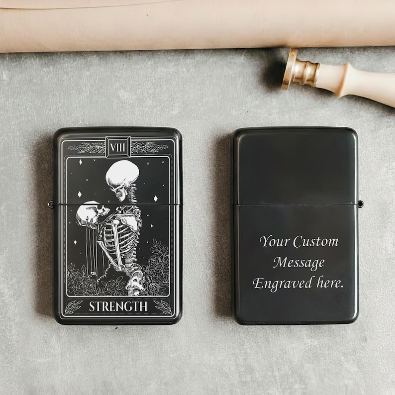 May include: Black metal lighter with a tarot card design featuring a skeleton figure and the word "STRENGTH". The other lighter is black with the text "Your Custom Message Engraved here."