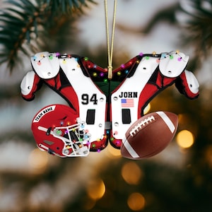 K&ouml;nnte beinhalten: Ein weihnachtlicher Football-Ornament. Es zeigt ein rot-wei&szlig;es Schulterpolster-Design, einen roten Helm mit dem Text "TEAM NAME" und einen braunen Football. Das Ornament enth&auml;lt die Nummer "94", den Namen "JOHN" und eine amerikanische Flagge.